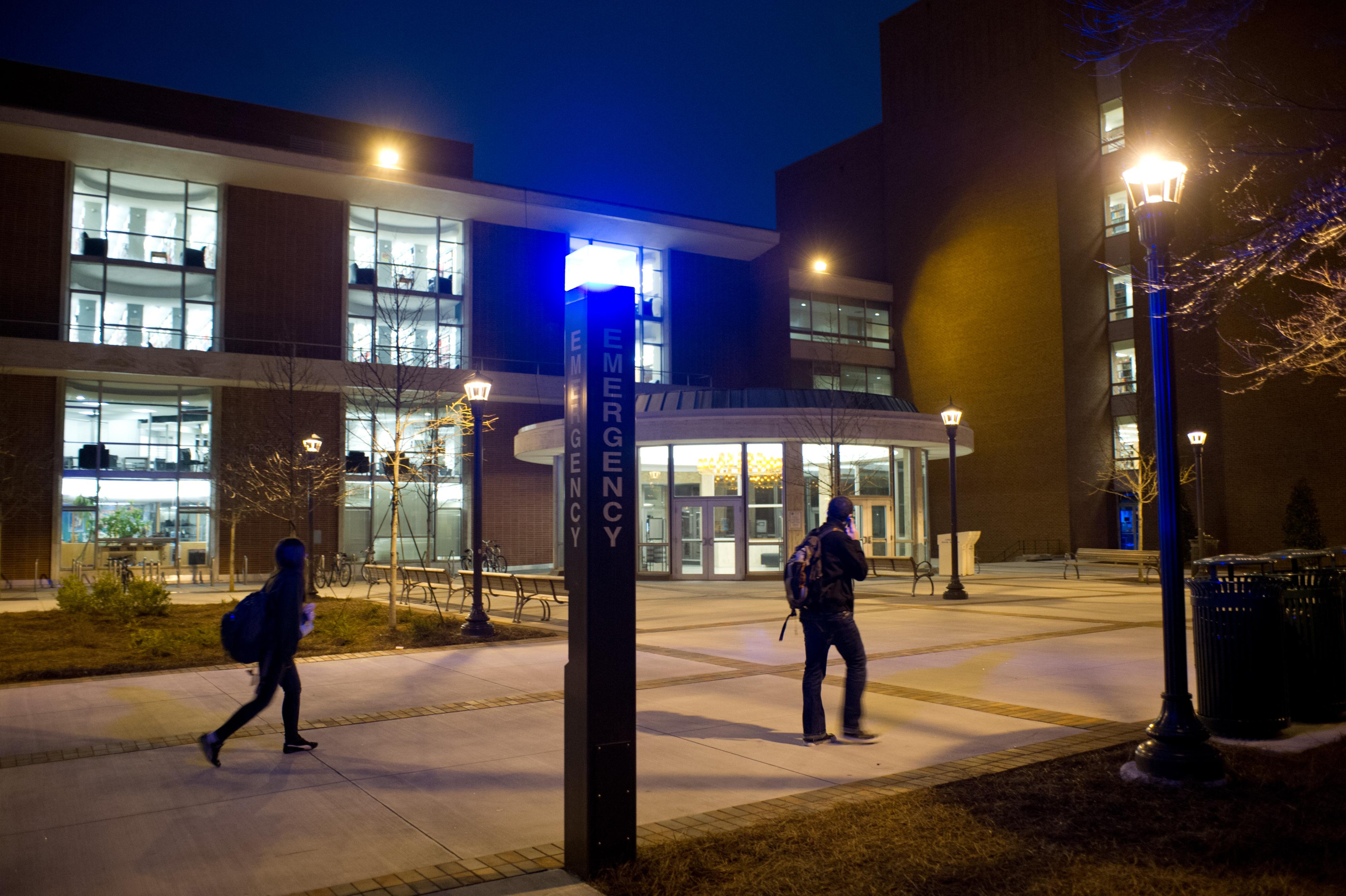 Exterior view of a campus emergency services building at night, featuring a tall illuminated "Emergency" sign near the entrance, with pedestrians walking across a plaza and building lights visible through large windows.
