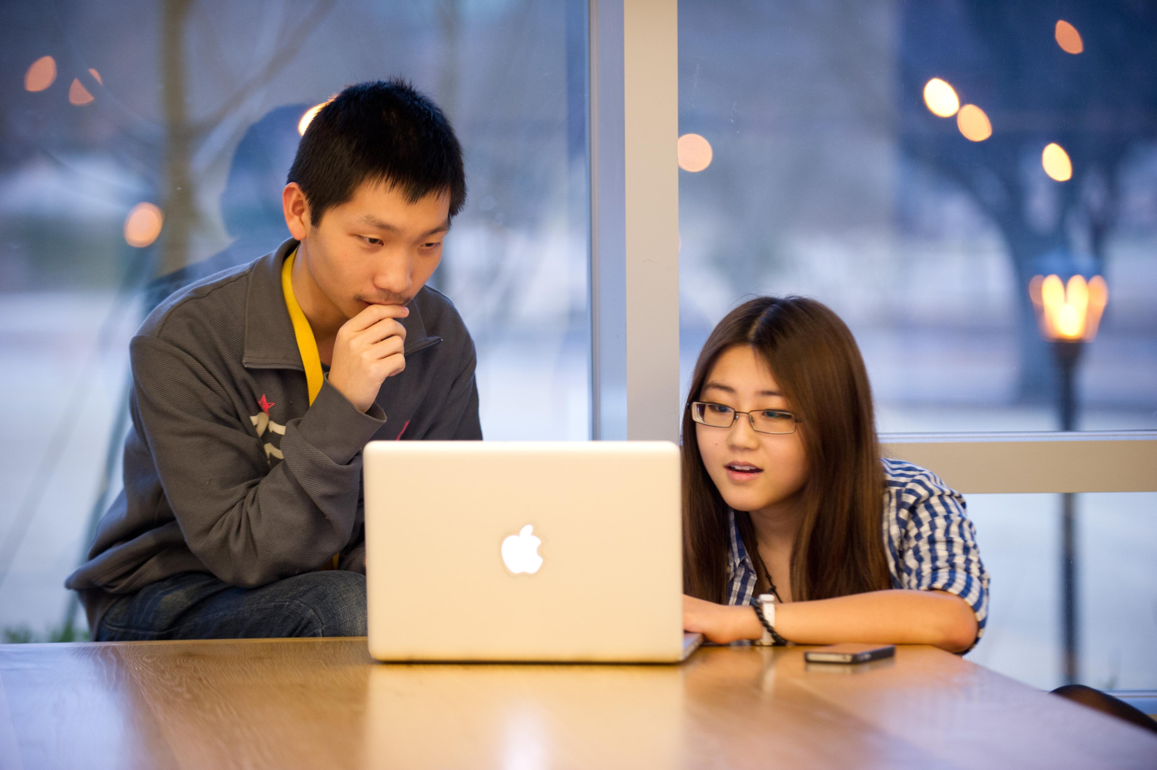 Two individuals collaborate on a laptop while seated at a table near large windows in a campus study space.
